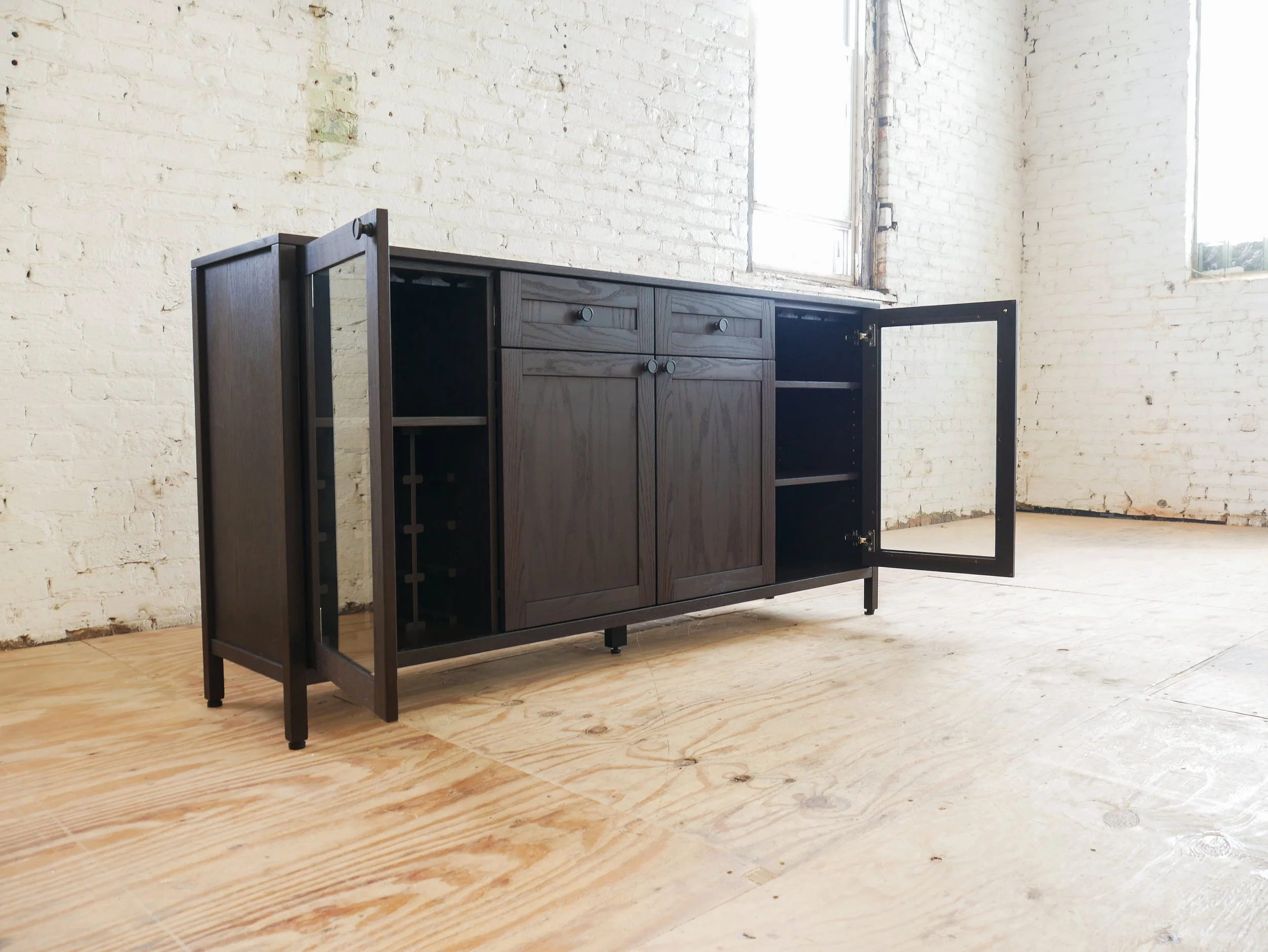 A black wooden cabinet with open doors and a drawer, standing against a white brick wall with two windows, on a wooden floor.