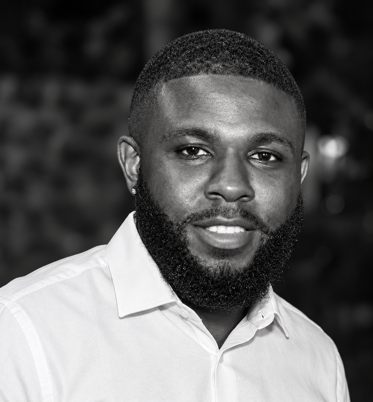 Black and white portrait of a young man with a short haircut and beard, wearing a white collared shirt, looking at the camera with a slight smile.