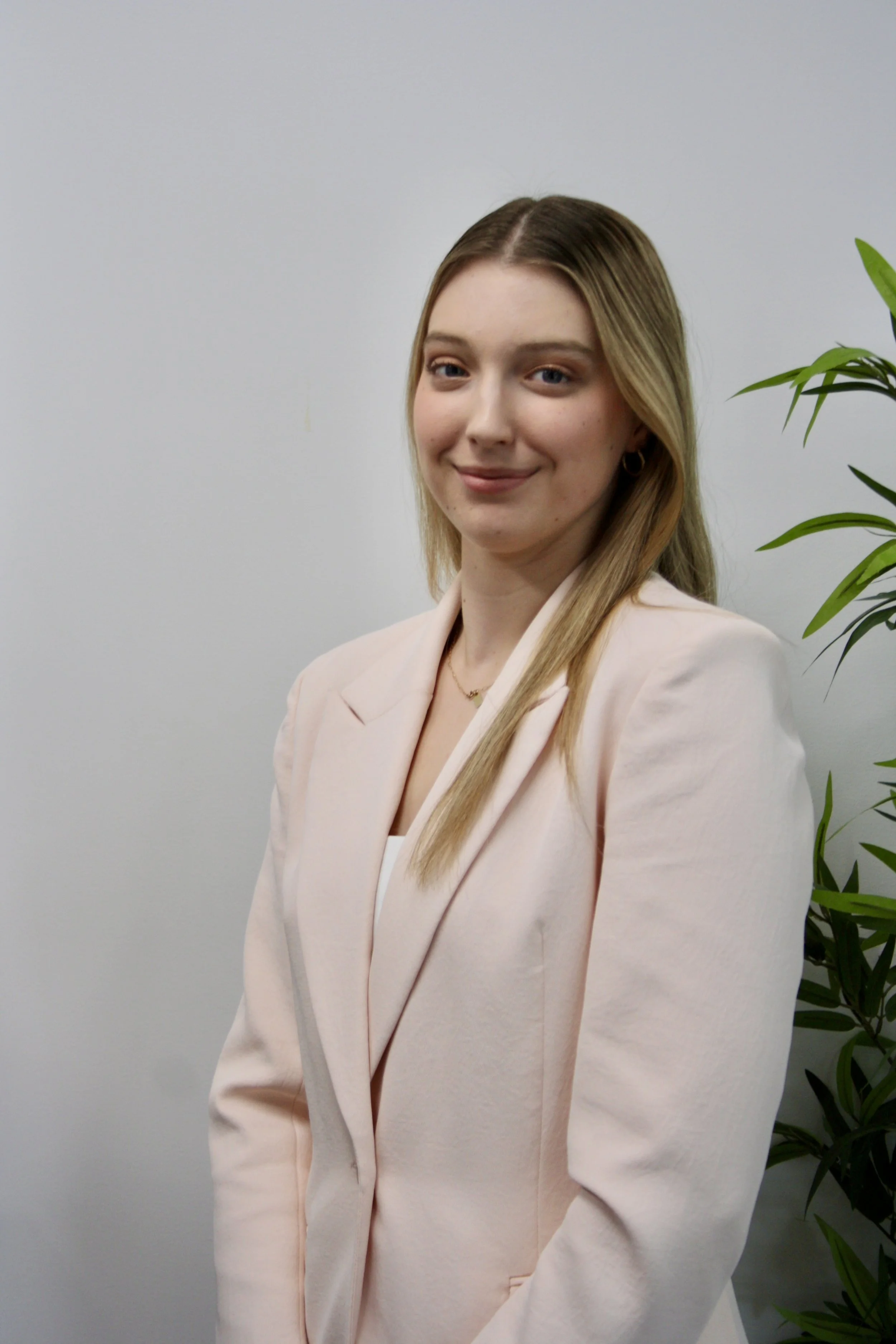 A woman with long blonde hair, wearing a light pink blazer and white top, standing indoors next to a green plant, smiling at the camera.