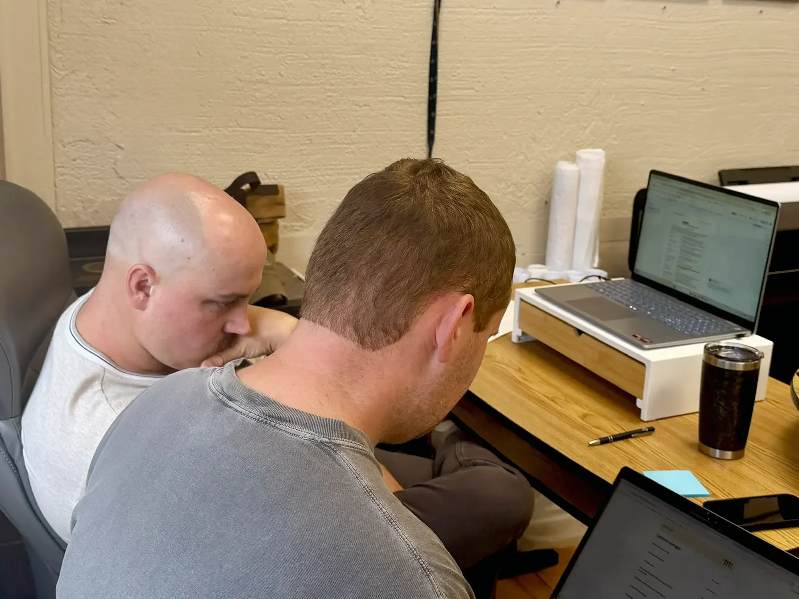 Two men sit at a desk looking at a laptop.