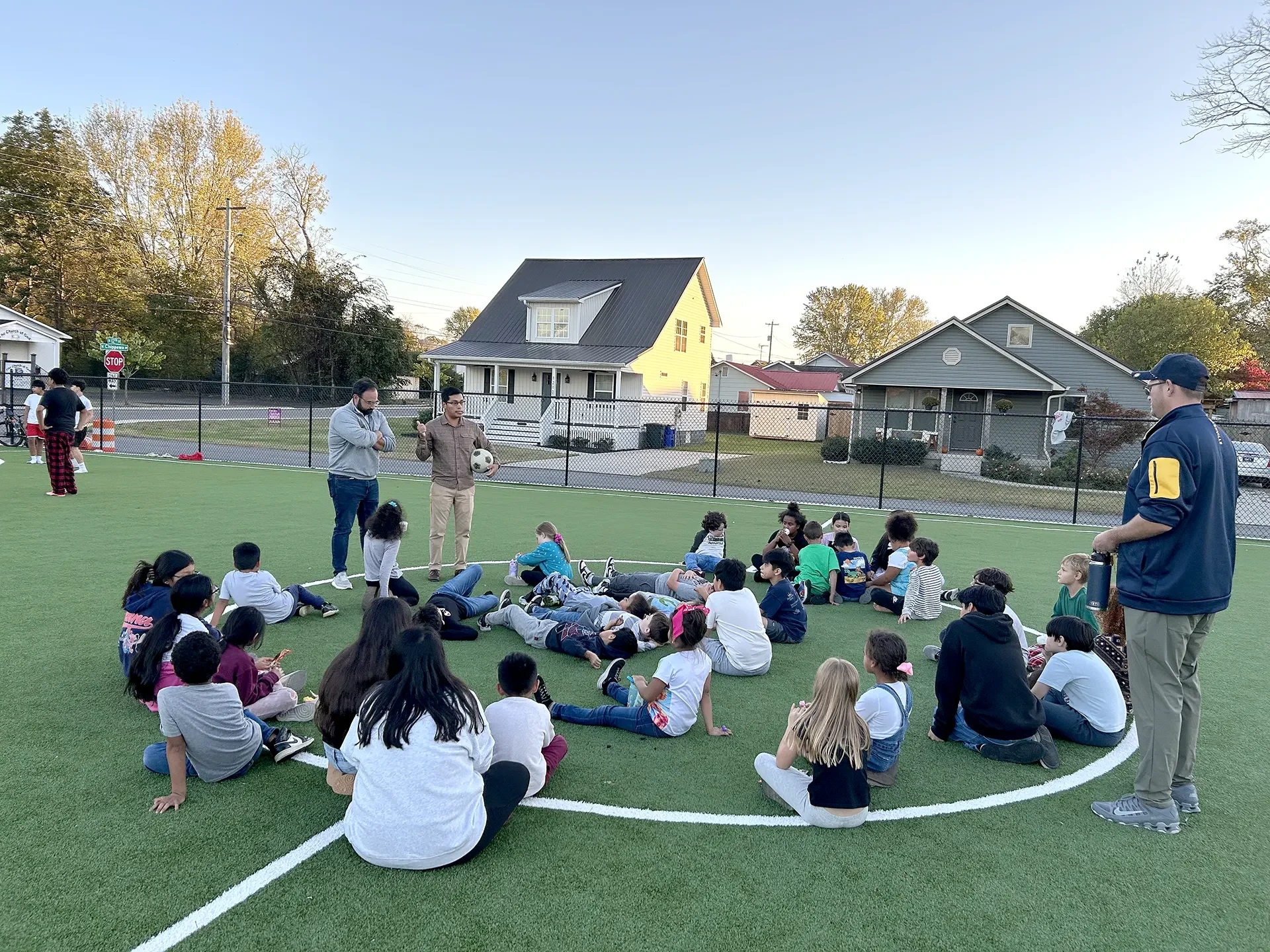 A group of kids sit on a soccer field listening to two coaches.