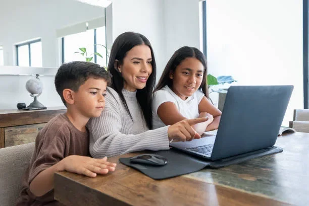 A woman and two children sit at a desk looking at a laptop.