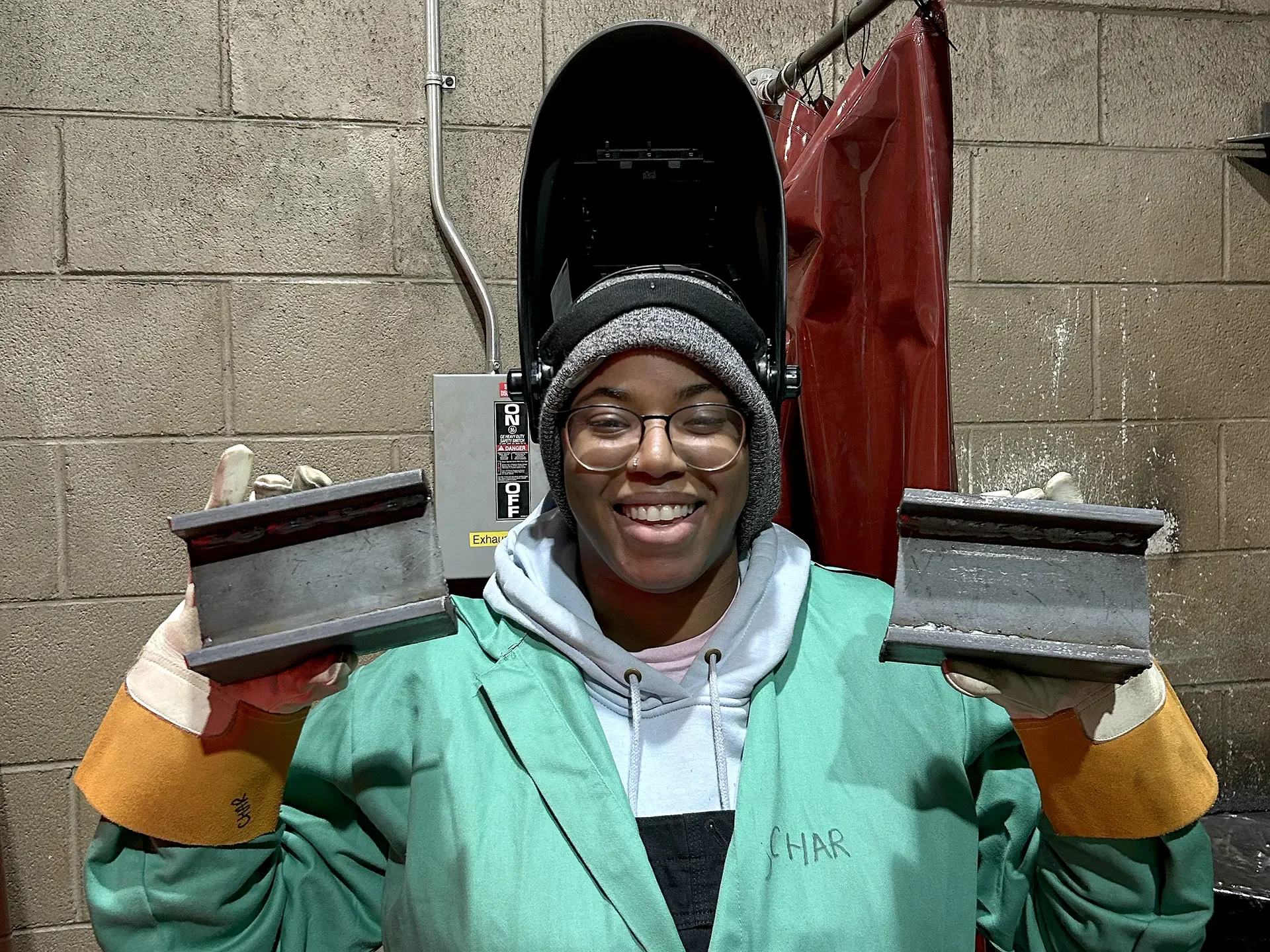 A woman wearing a welding visor holds up a metal bar that has been cut.