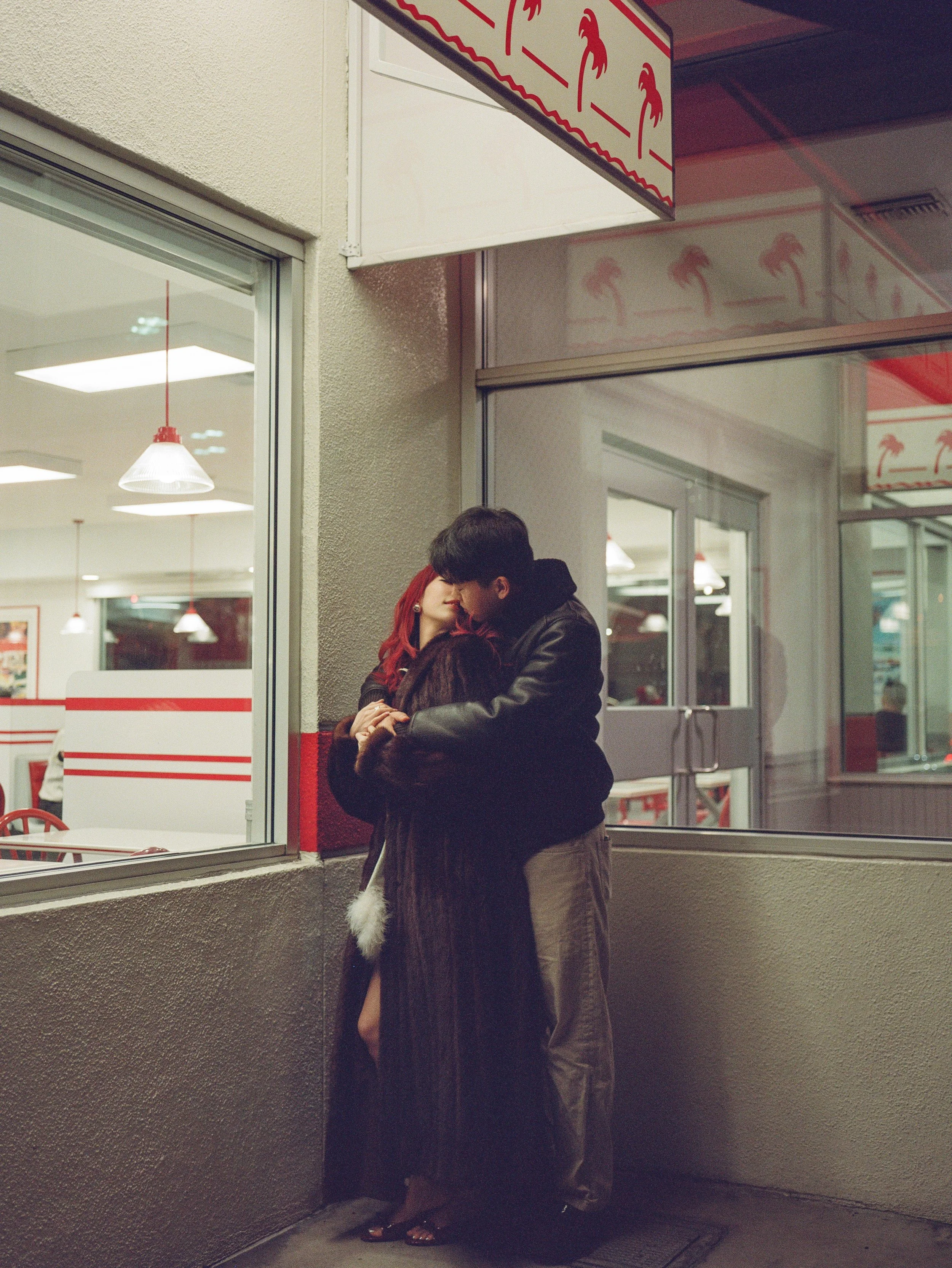 A couple sharing a kiss outside a building with glass windows reflecting a red and beige sign featuring palm trees. The woman has long red hair, and the man is wearing a black leather jacket. They are embracing each other near the corner of the build