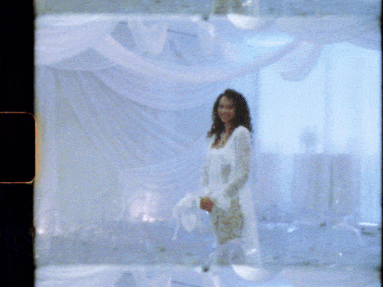 A woman in a white outfit stands in front of a decorated backdrop, holding a garment, at an indoor event.