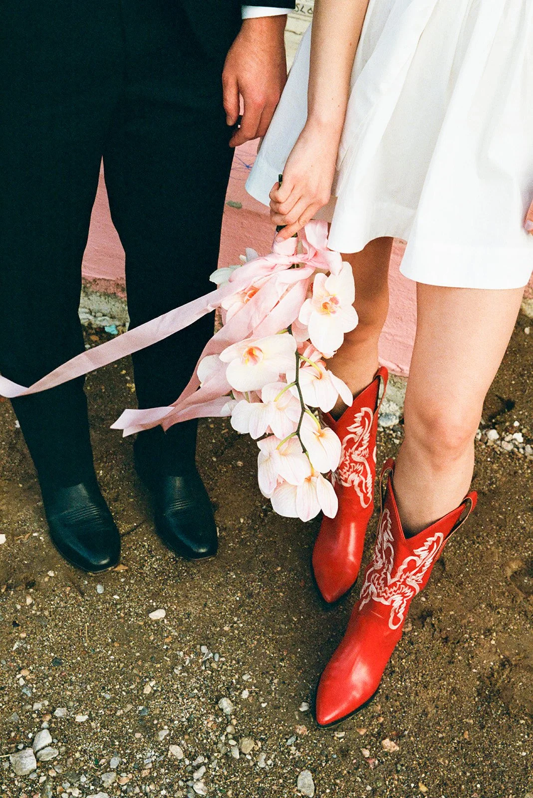 Close-up of a couple holding a bouquet of pink orchids, with the woman wearing red cowboy boots and a white dress, and the man wearing black shoes and black pants, on a dirt ground.