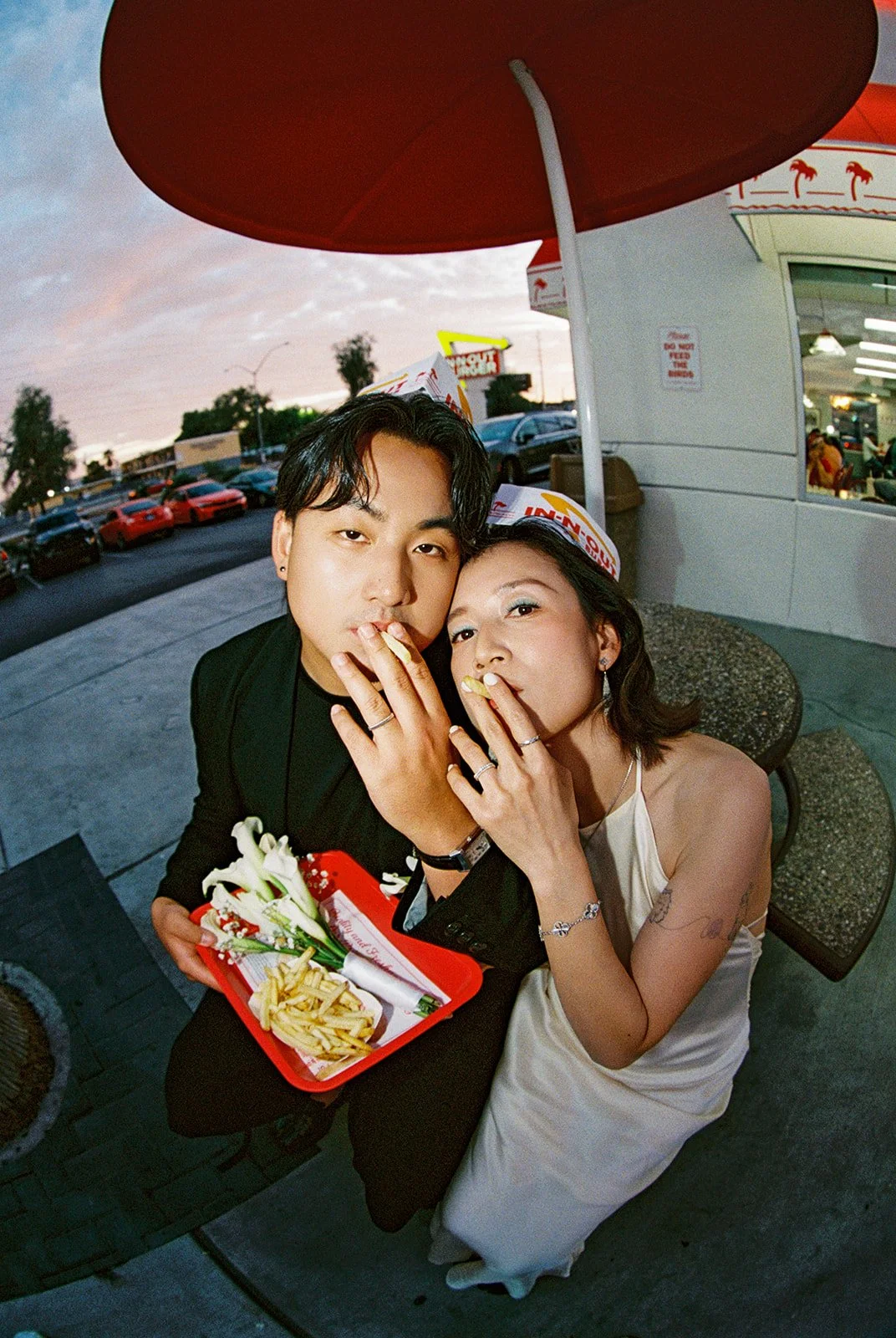 A young man and woman are sitting together outside a restaurant at sunset, sharing fries and sushi, wearing paper hats from In-N-Out Burger, with a parking lot and sunset sky in the background.