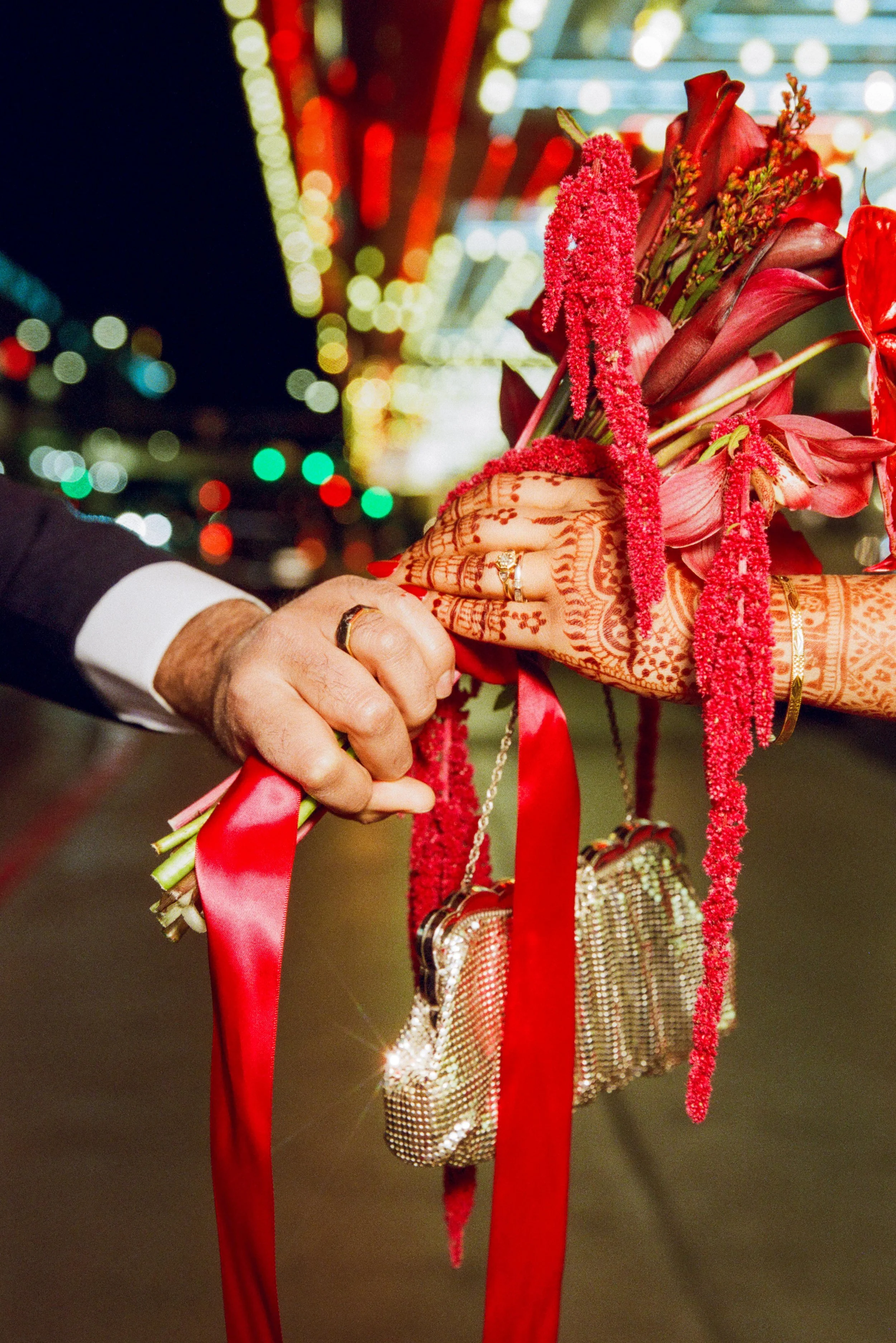 Close-up of a woman’s henna-decorated hand holding a bouquet with red flowers and ribbons. In the background, colorful, blurred lights create a festive atmosphere.