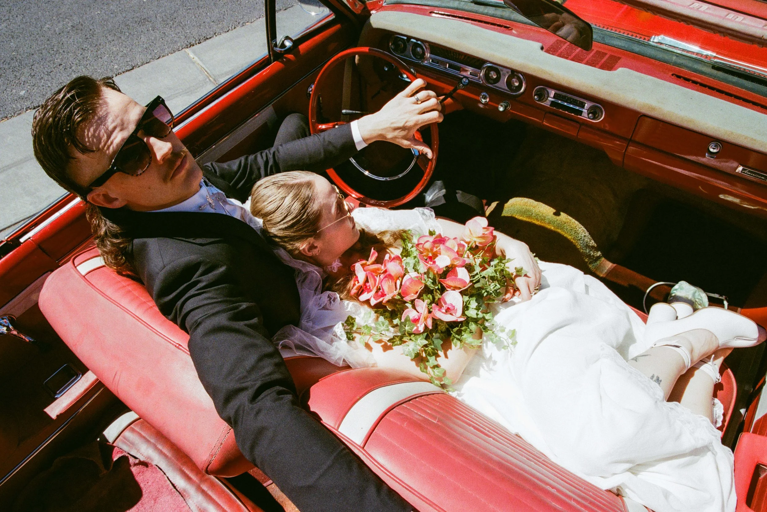 A man in sunglasses and a dark suit is driving a vintage red convertible with a woman in a white wedding dress and veil sitting beside him, holding a bouquet of pink flowers. They are on a street with sunlight casting shadows inside the car.