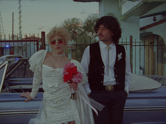 Two people standing next to a vintage car, with a girl holding a bouquet of flowers and wearing a white dress, and a guy in dark clothing with sunglasses.