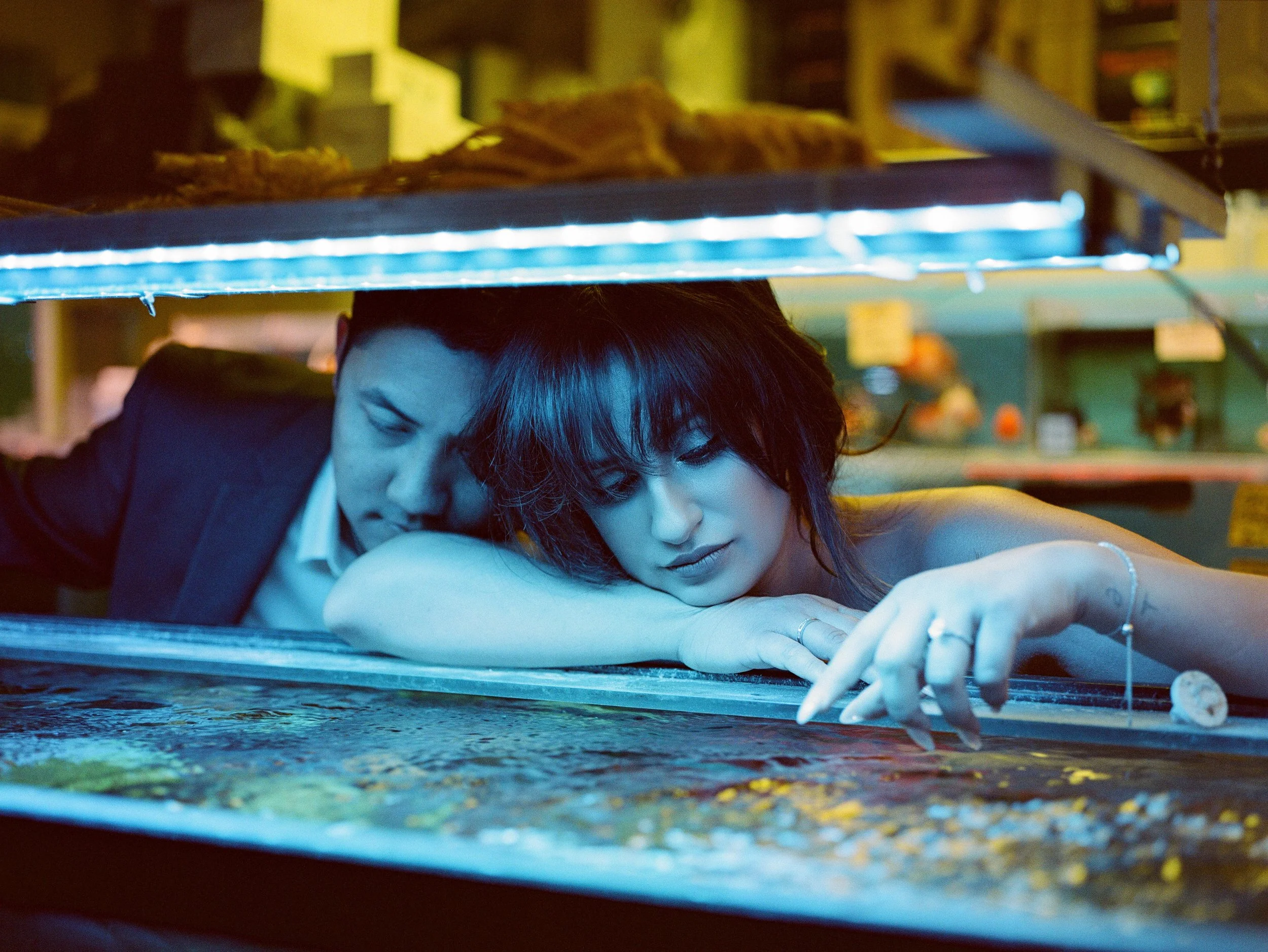 A woman with dark hair leaning over a table or counter, reaching towards a surface below her. The setting appears to be industrial or workshop-like, with shelves and various objects in the background. The lighting is dim with a blue tone.