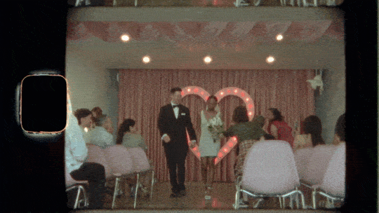 A person in a tuxedo walking in front of seated guests at a wedding reception with a heart-shaped backdrop decorated with lights.