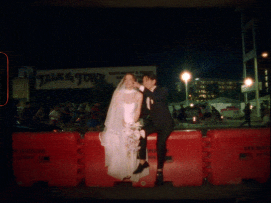 A bride and groom standing close together at night, holding hands and gazing into each other's eyes, with a row of barricades and parked cars in the background.