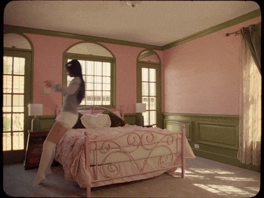 A woman in a pink and green bedroom with large windows, standing next to the bed, holding an object.
