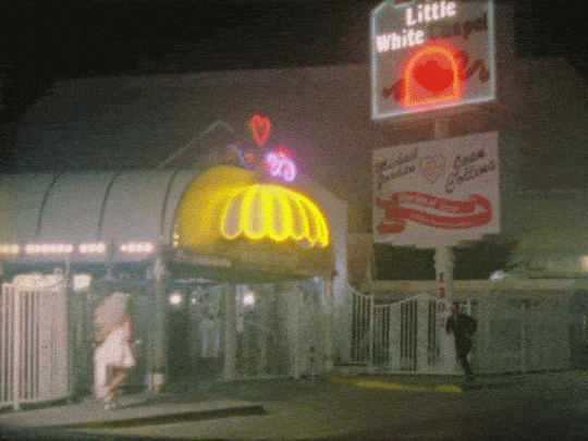 Nighttime scene with a brightly lit storefront, neon signs, and two people walking by, one wearing a hat and the other carrying a bag.
