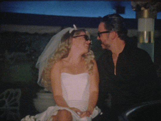 A woman dressed as a bride and a man dressed as a groom sitting closely together, smiling at each other during a wedding celebration.