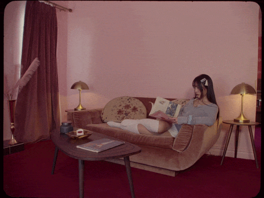 Young woman in a gray sweater and white skirt sitting on a beige sofa, reading a magazine in a cozy living room with pink walls, red carpet, and gold lamps.
