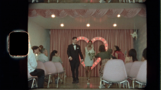 A wedding ceremony taking place on a stage with a pink curtain backdrop, decorated with a large illuminated heart. The bride and groom are walking down the aisle, with guests seated on both sides.