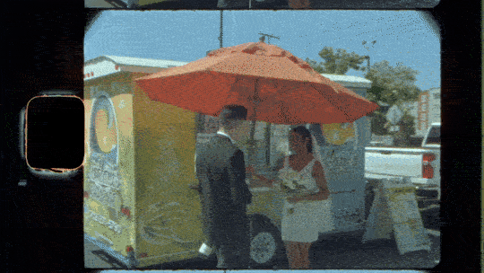 A man in a suit and a woman in a white dress talking under an orange umbrella outside a food truck.
