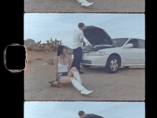 A woman sitting on the ground next to a white convertible car with its hood open, while a man stands nearby looking under the hood in a desert landscape.