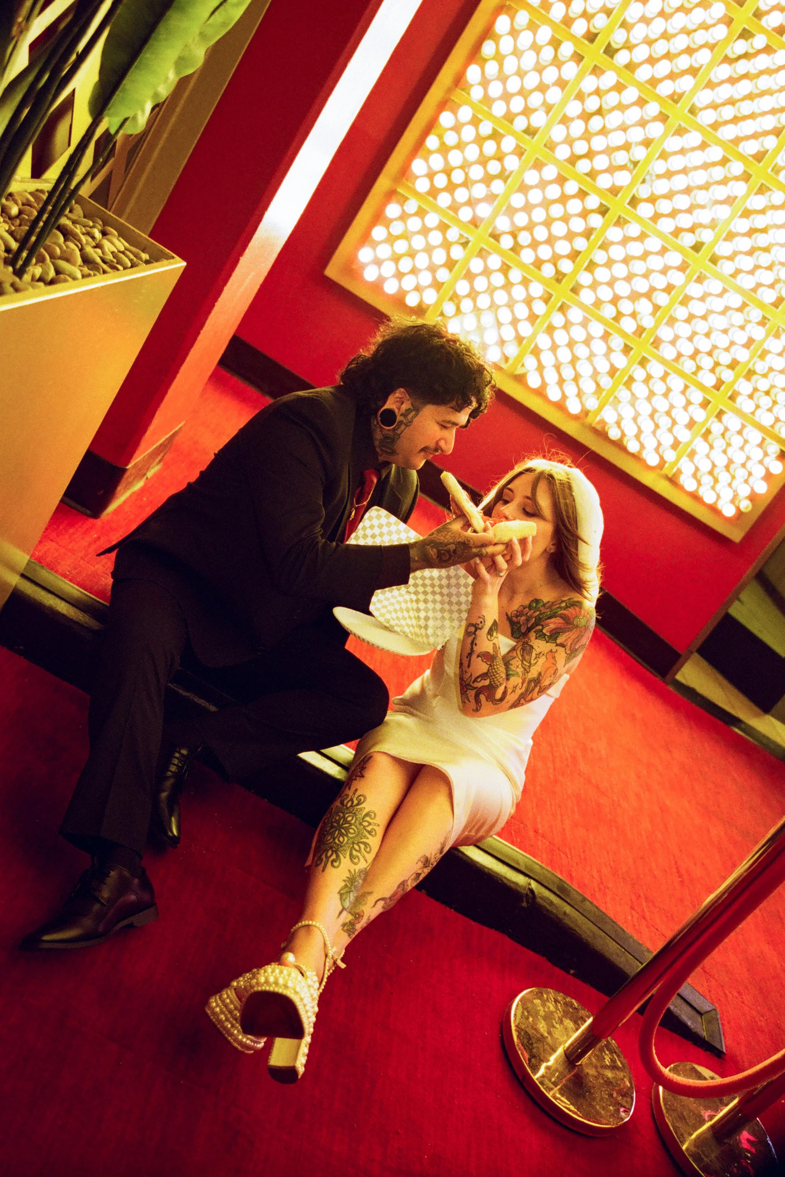 A man with tattooed face and neck, dressed in a black suit, is holding a sandwich for a woman with tattoos on arms and legs, who is sitting on the floor in a white dress and gold platform sandals in a red-lit room with a decorative light fixture on the wall and plants in the background.