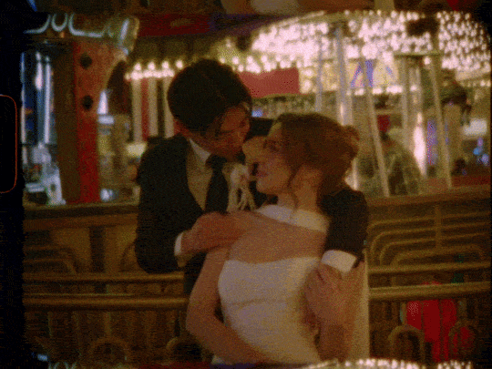 A man and woman are sharing a romantic moment in a dimly lit restaurant or bar, with decorative lighting and a festive atmosphere.