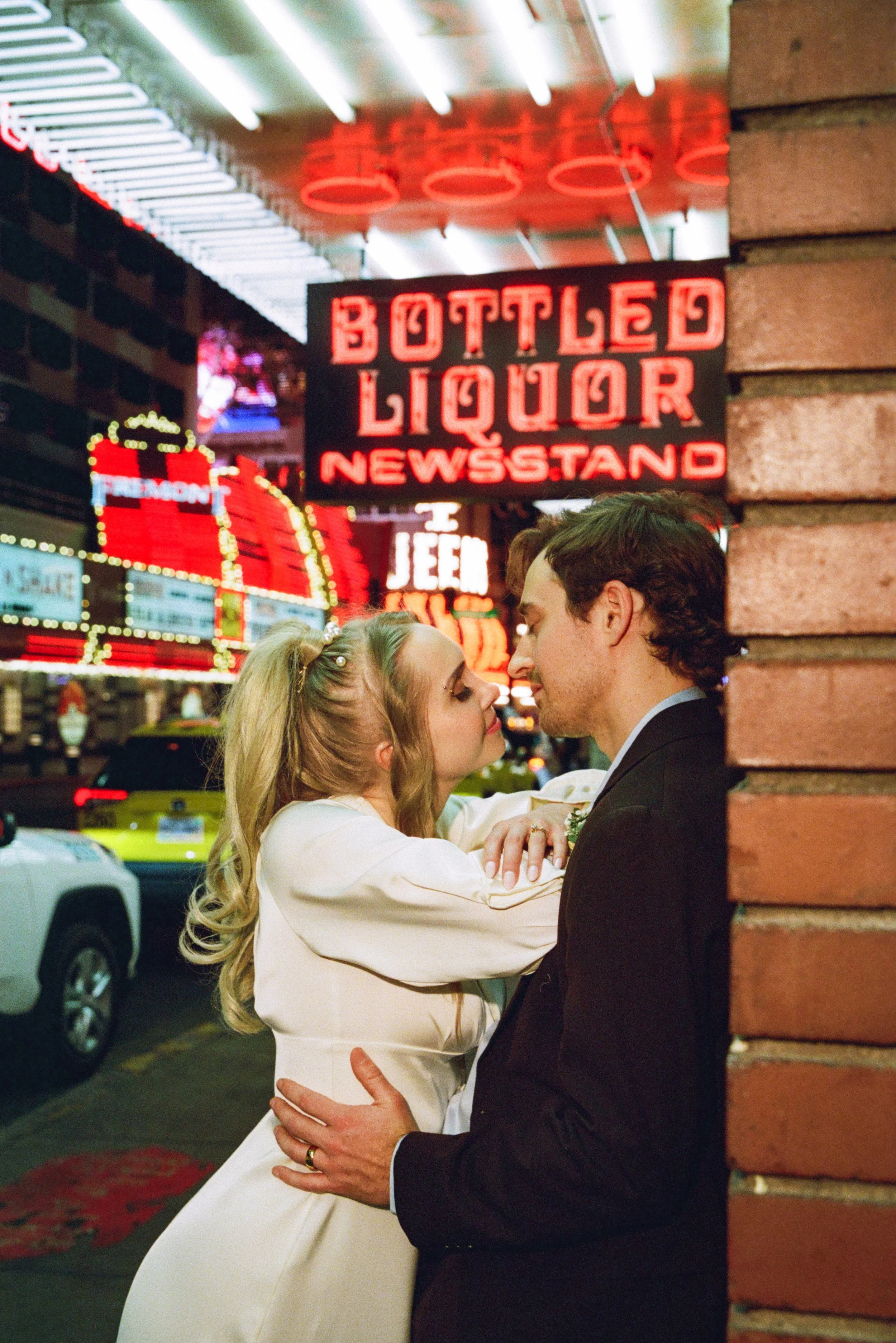 A couple dressed in wedding attire are standing close together on a city street at night, partially obscured by a brick wall. Neon signs and busy traffic are visible in the background.