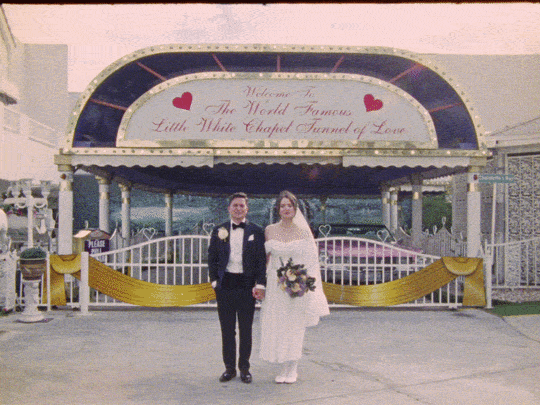 A couple in wedding attire standing in front of a decorated entrance with an arch that has a sign reading 'Welcome to The World Famous Little White Chapel Where Love Begins.'