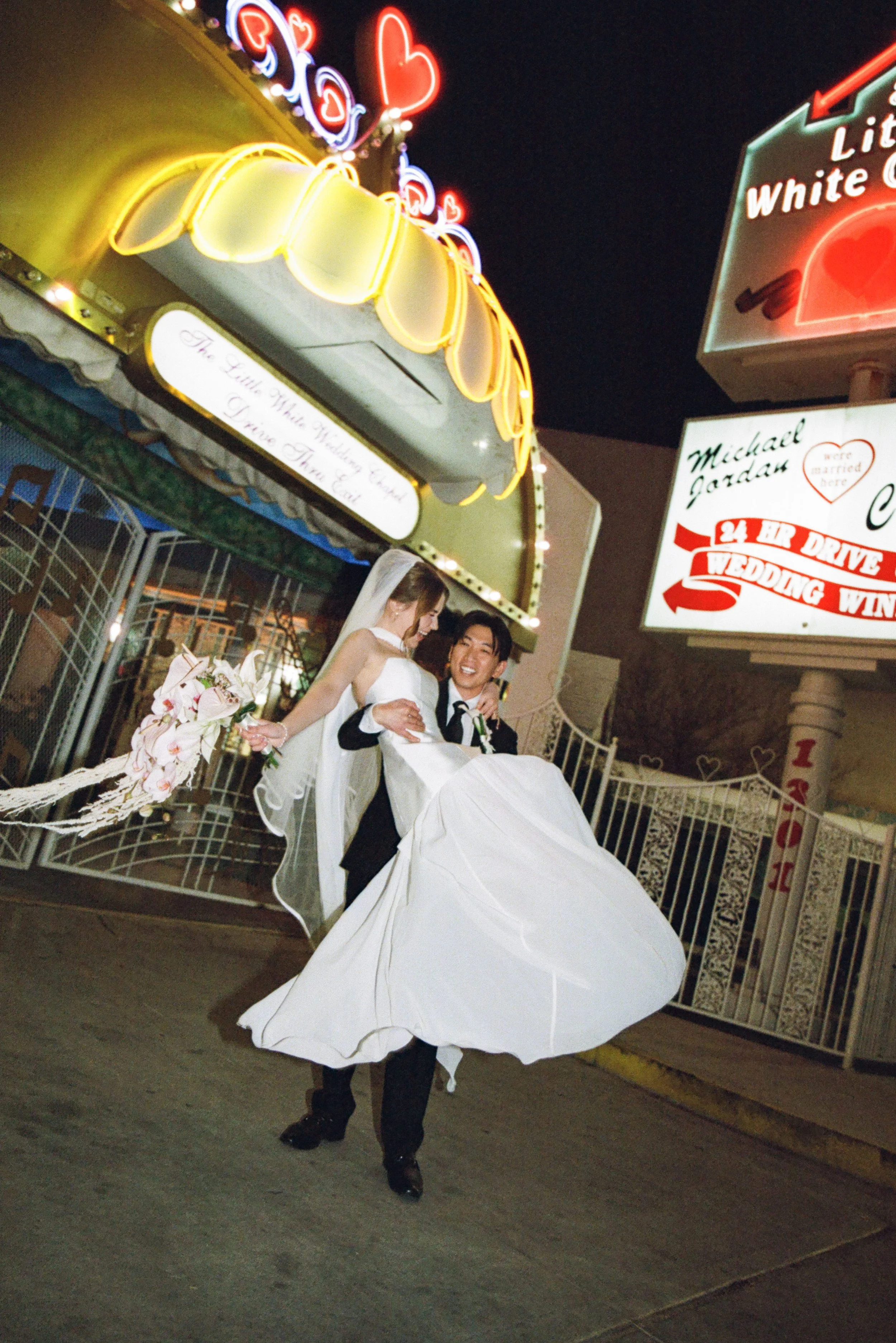 A newlywed couple sharing a joyful moment under neon signs and illuminated signs at night, with a bride in a white wedding gown and veil, and a groom dressed in a black tuxedo. The groom is lifting the bride while she holds a bouquet of flowers.