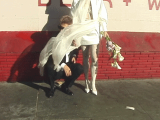 A person is crouching beside a white statue of a woman holding flowers, with a red brick building wall in the background.