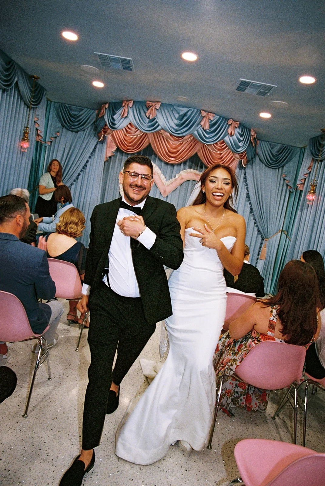 A bride and groom at their wedding reception. The groom in a tuxedo and the bride in a white wedding dress are smiling and posing for the camera. Guests are seated at tables around them, and the room is decorated with blue and pink drapes and curtain
