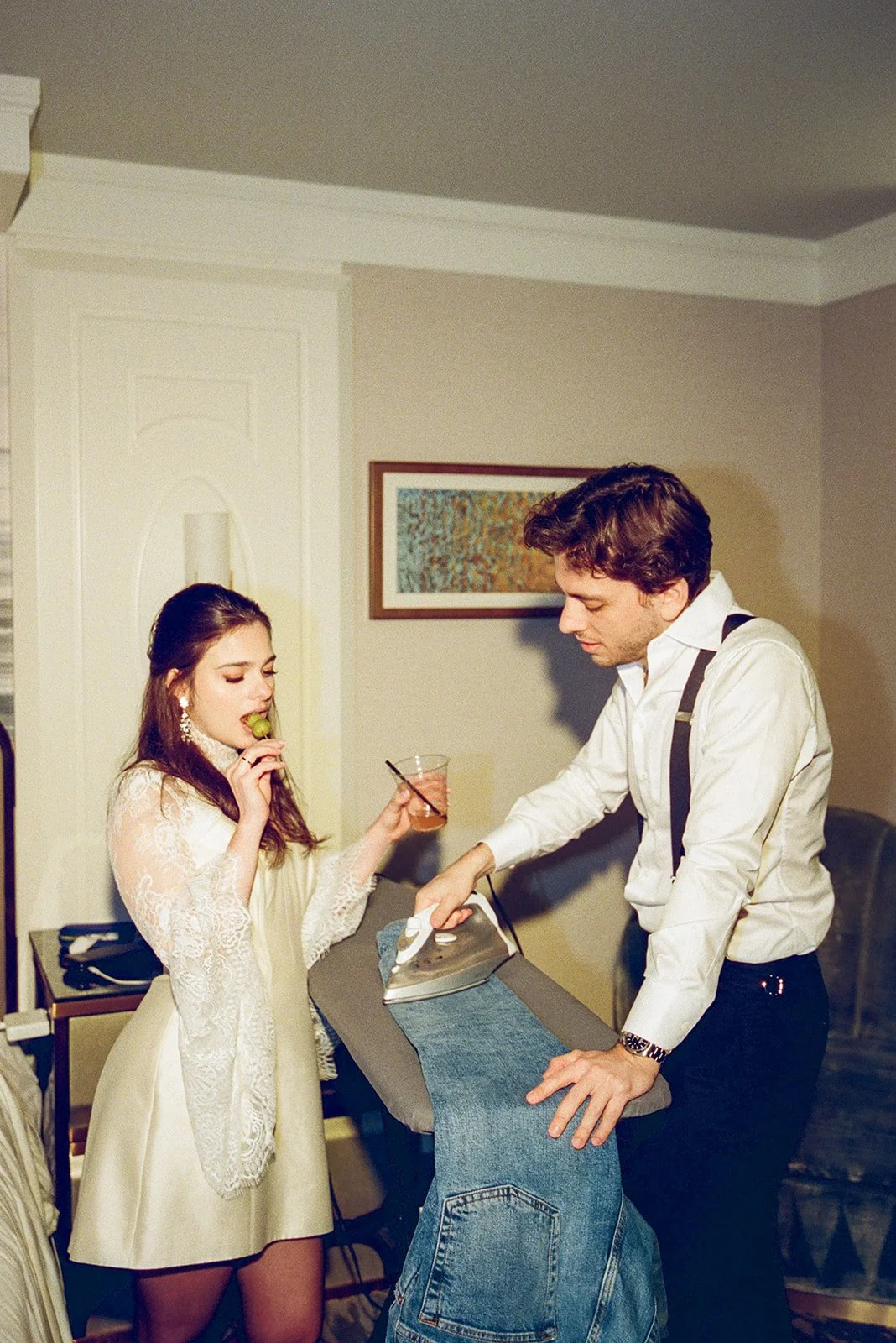 A young woman with long brown hair, wearing a white lace dress, holds a glass of drink and a snack, standing next to a man in a white shirt with suspenders, who is ironing a pair of jeans.