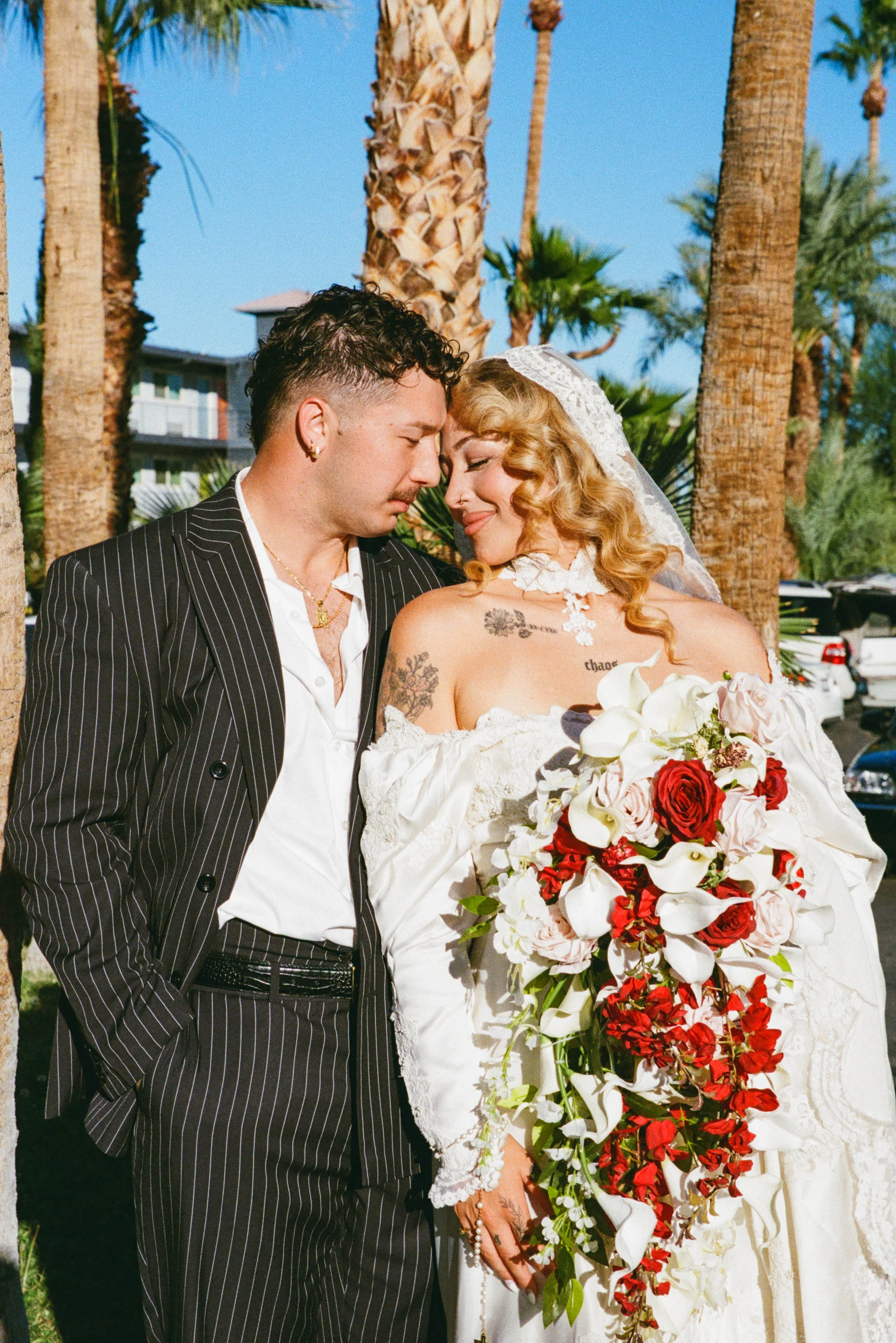 A bride and groom sharing a tender moment outdoors, with palm trees in the background, the groom in a black pinstripe suit and the bride in a white wedding dress holding a large bouquet of white and red roses.