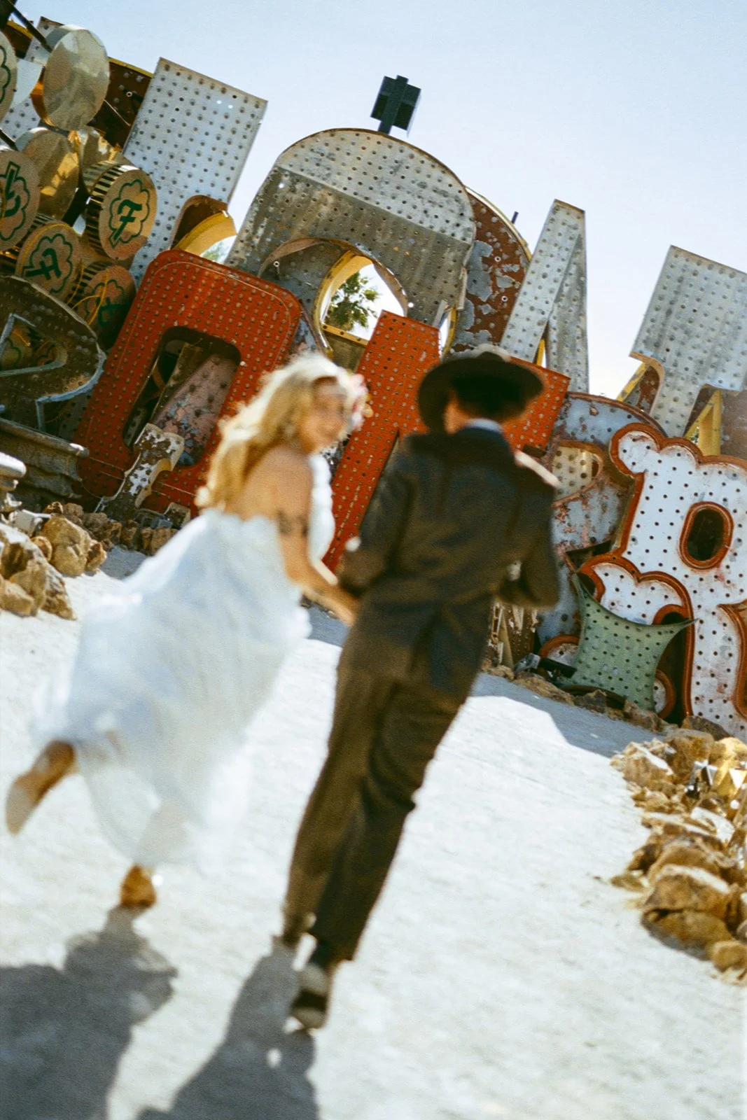 A couple dressed in vintage-style clothing walking hand in hand in a desert area with large, weathered neon signs in the background.