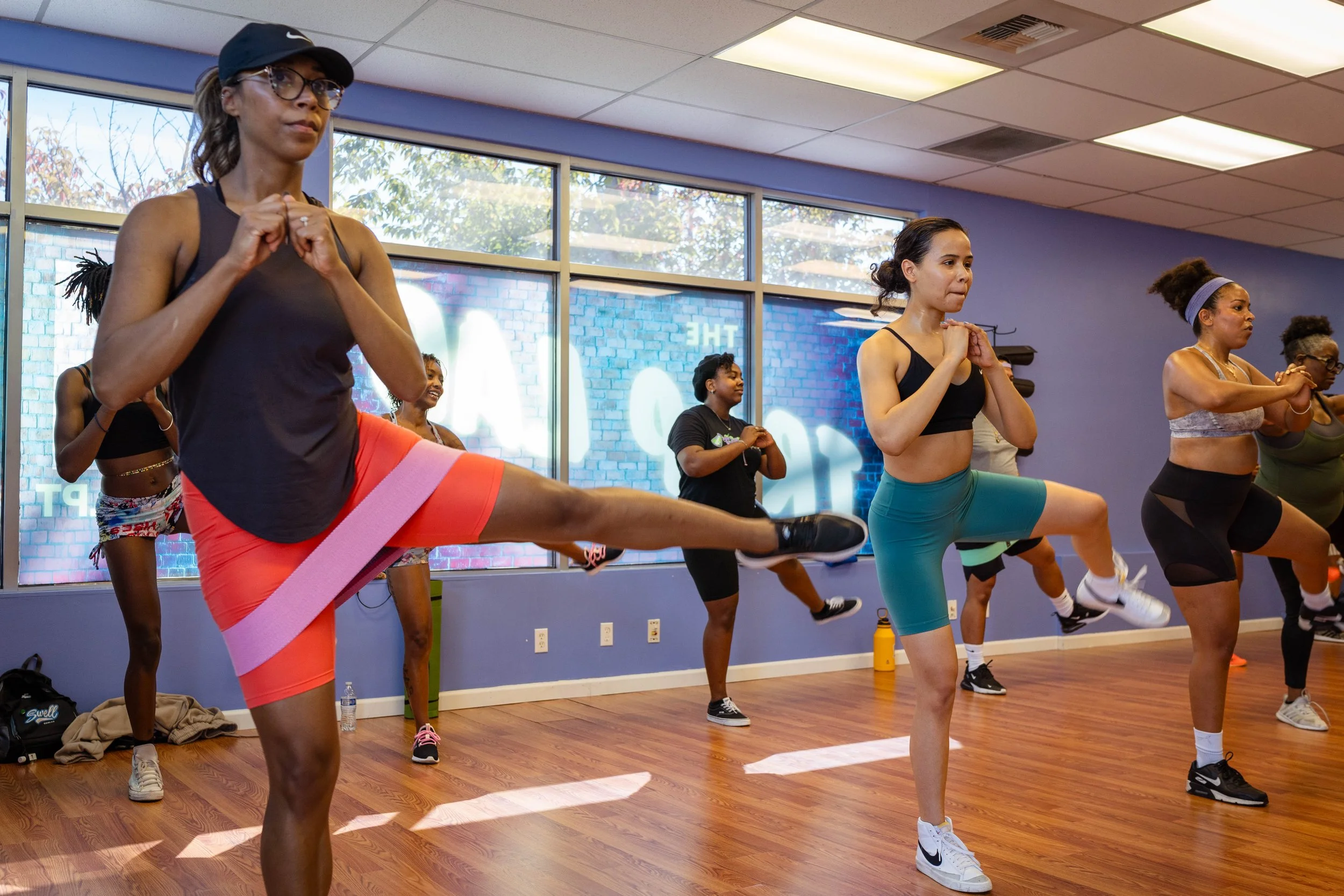 Group of women participating in a fitness class doing leg lifts on a wooden floor in a room with large windows and purple walls.