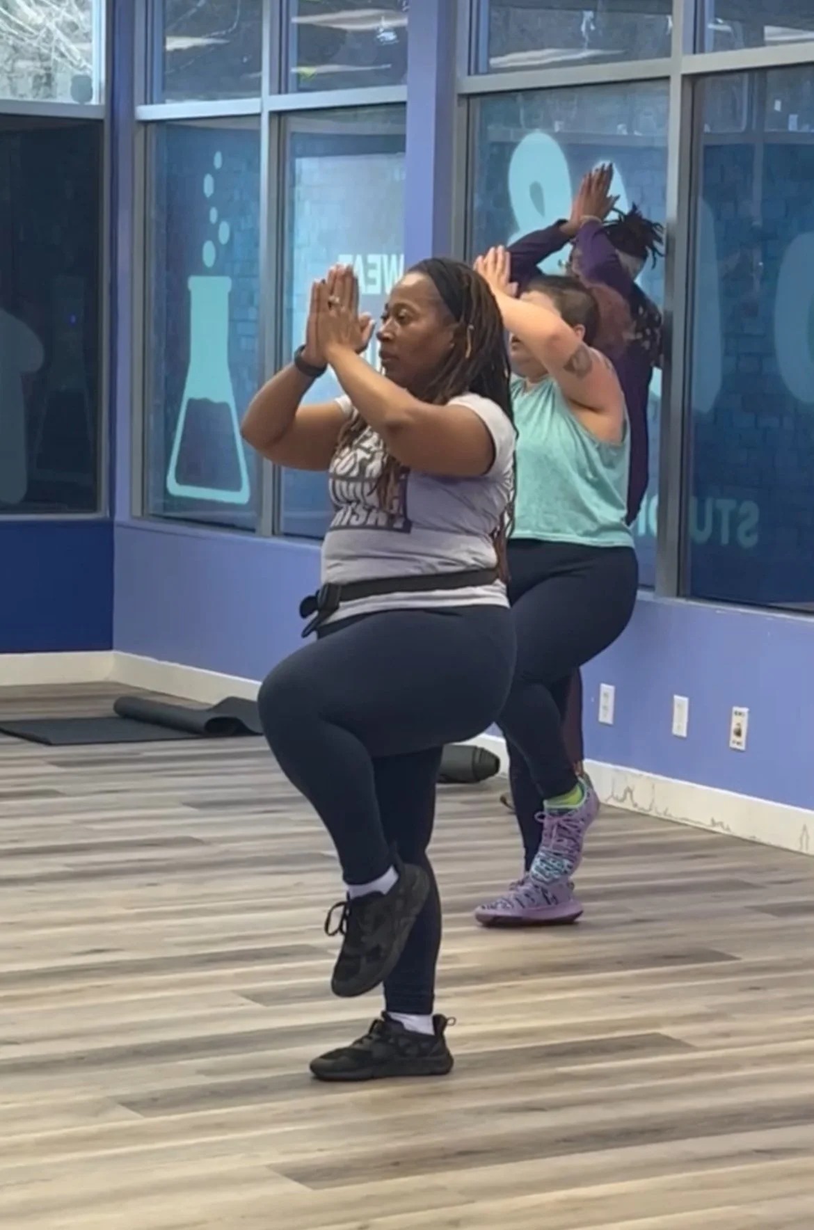 Two women participating in a fitness class at a gym, balancing on one leg with hands pressed together in front of their chests, practicing yoga or balance exercises.