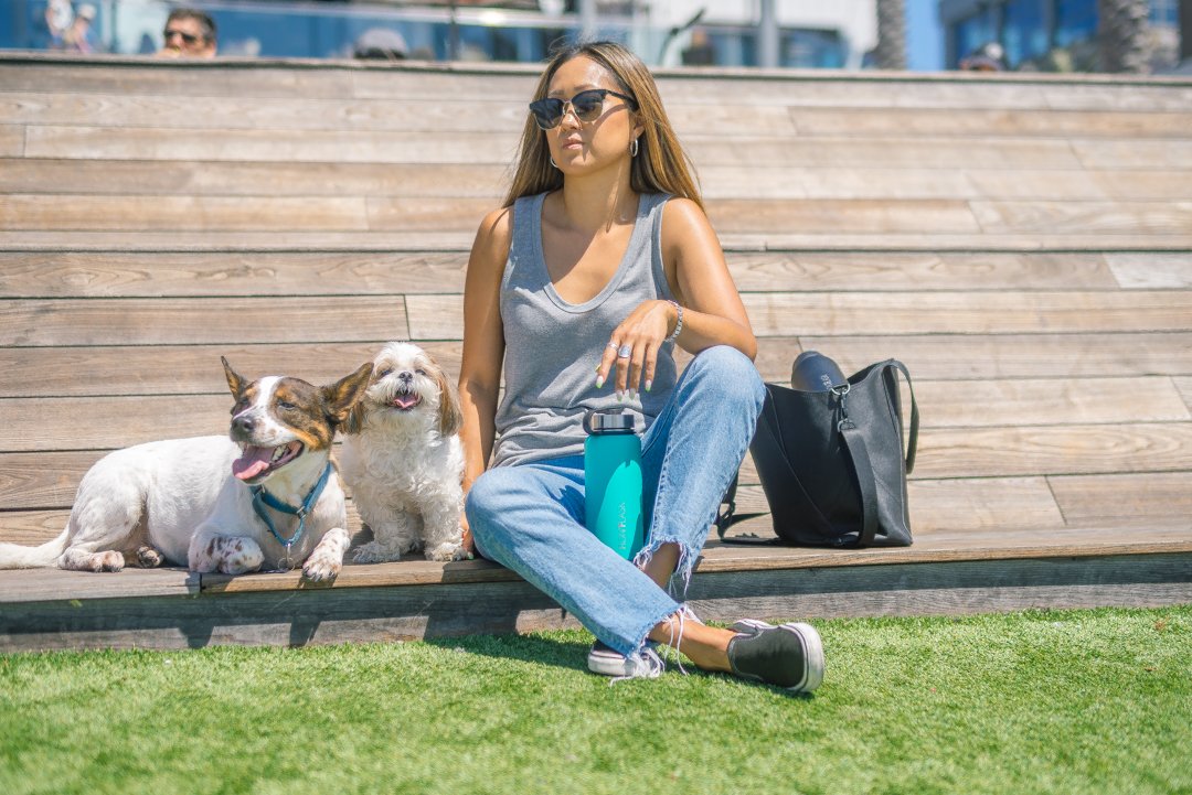 Woman sitting on wooden steps with two dogs, wearing sunglasses and casual clothing, in an outdoor setting with a black bag and a blue water bottle.