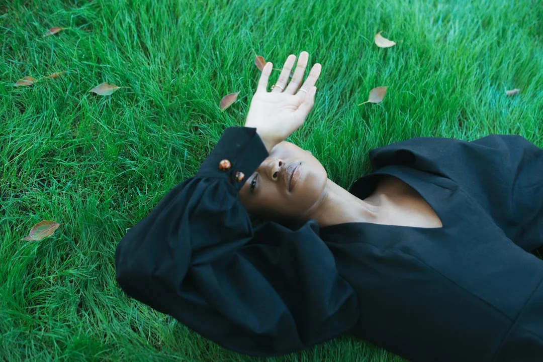 A woman lying on green grass with her eyes closed, wearing a black jacket, and holding her right hand above her face in a relaxed pose.