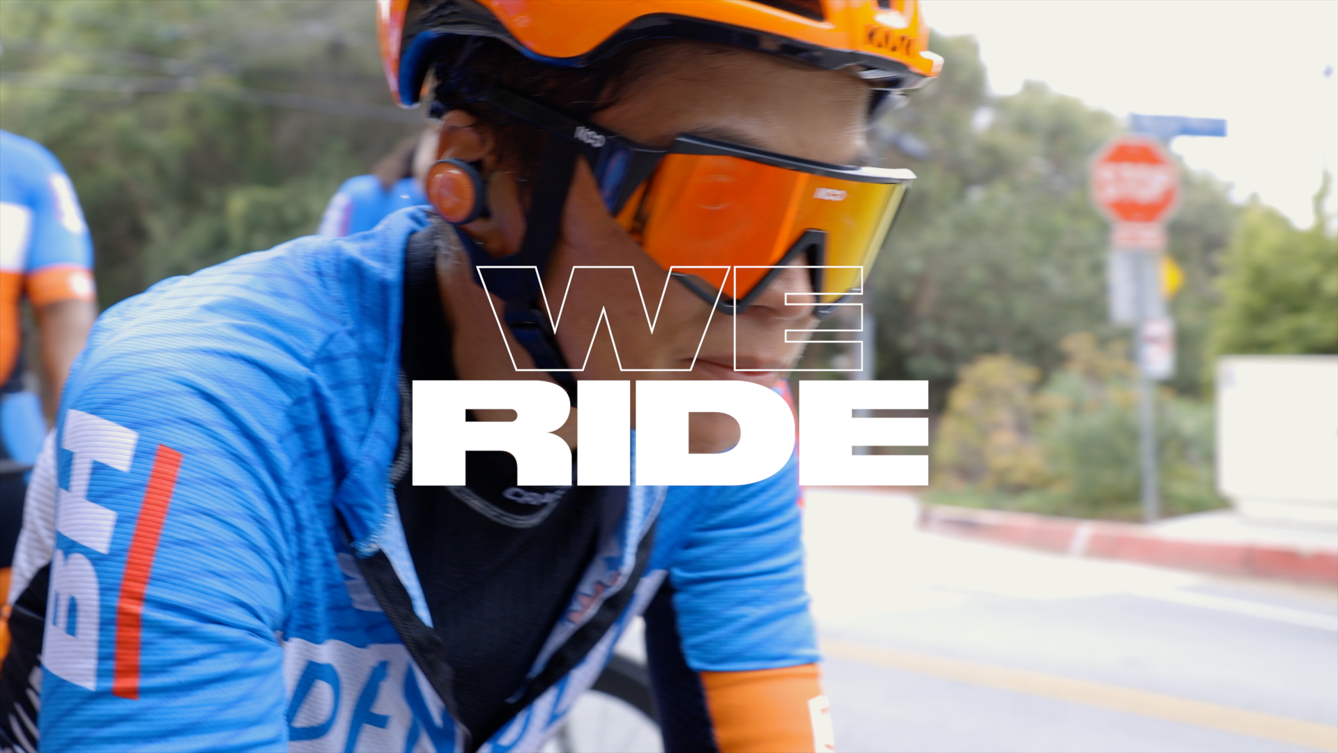 A cyclist wearing an orange helmet and sunglasses on a blue jersey riding during daytime, with a background of trees, street signs, and a clear sky, overlaid with the text 'WE RIDE'.