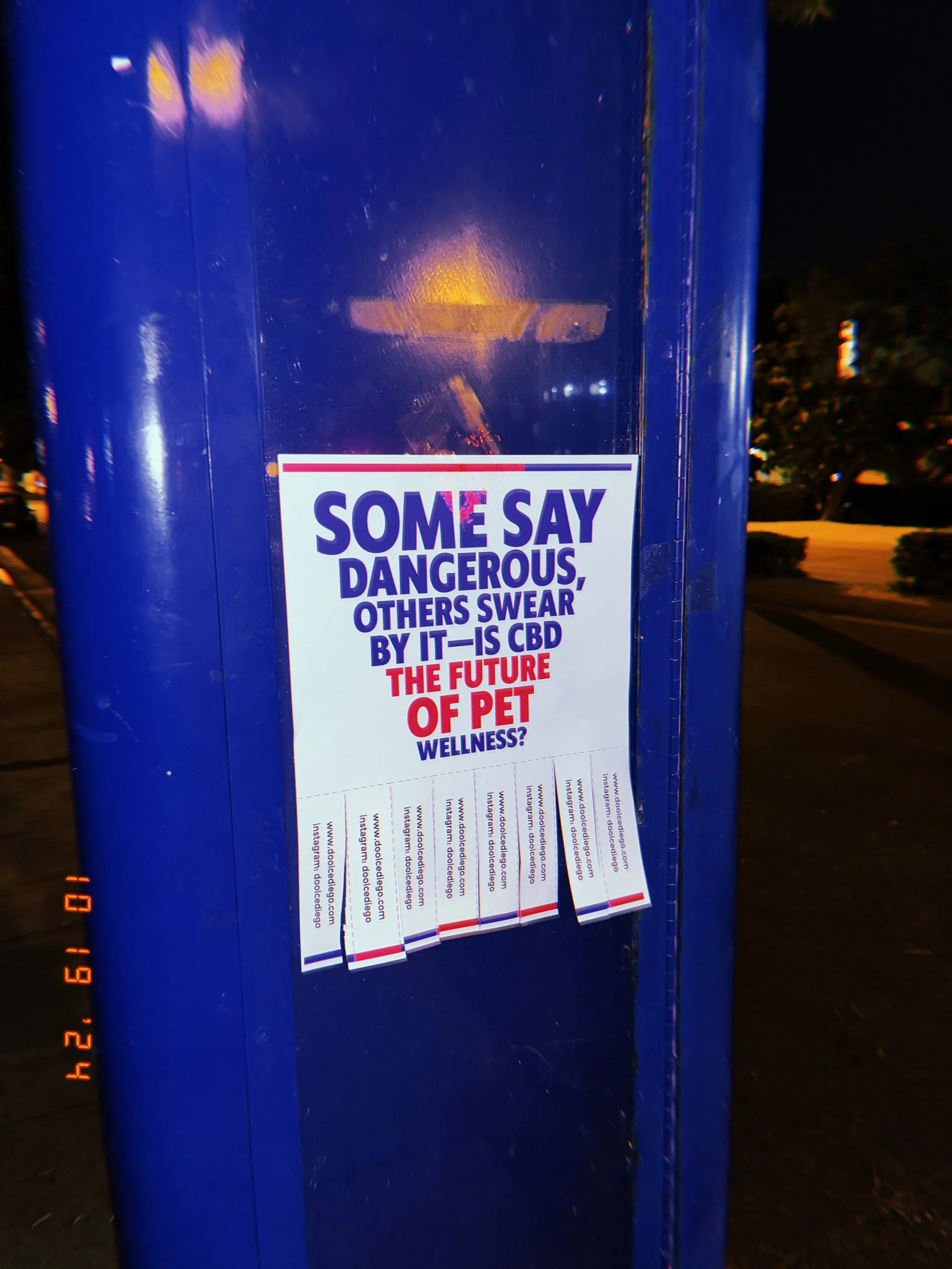 A poster on a blue pole at night with the text: 'Some say dangerous, others swear by it— is CBD the future of pet wellness?' and social media information at the bottom.