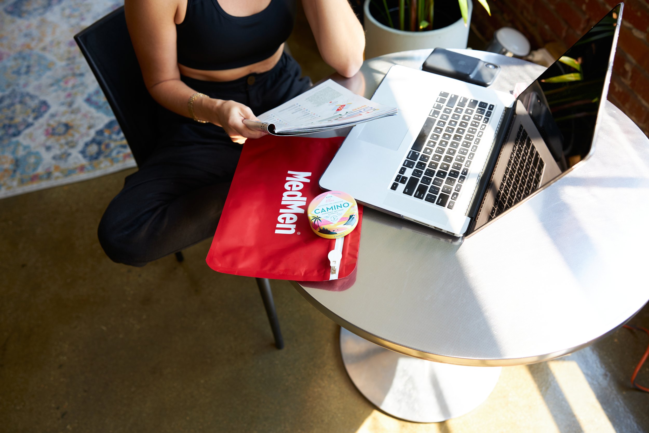 Person sitting at a table using a laptop with papers in hand. On the table, there is a red MegMent bag and Camilo lip balm, with a smartphone and a large plant in the background.
