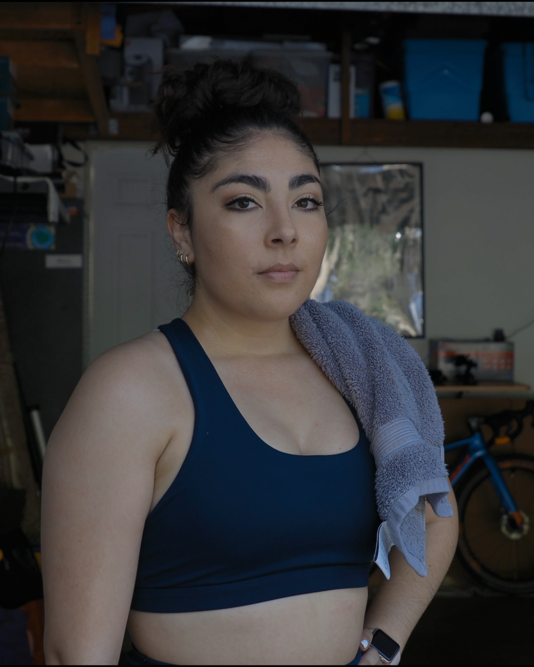 A young woman standing in a garage, wearing a navy sports bra with a gray towel draped over her shoulder, and an Apple Watch on her wrist, with gym equipment and a bicycle in the background.