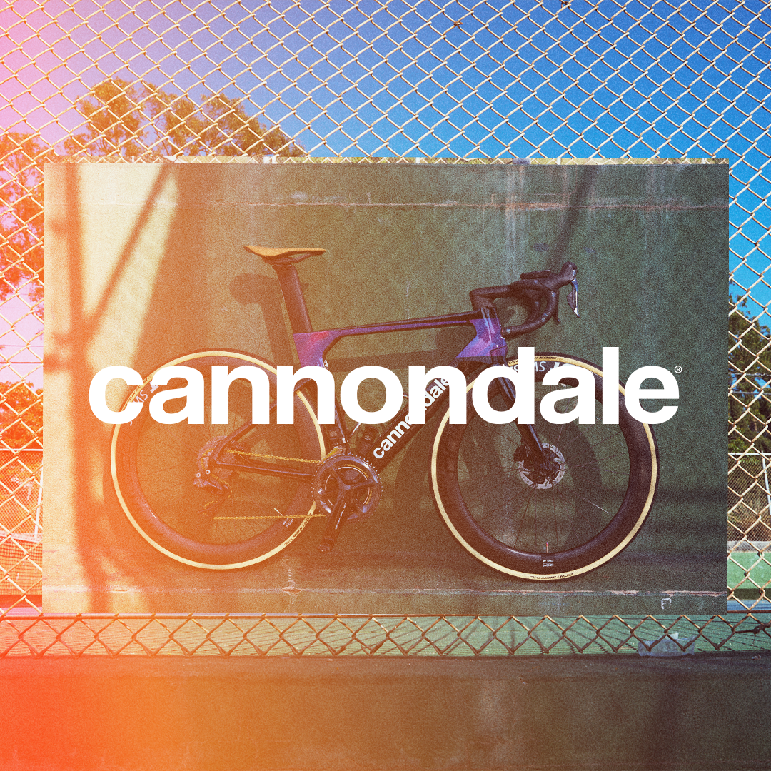 A Cannondale road bicycle with black and purple frame, yellow tires, and a yellow seat, displayed against a green background with a chain-link fence and trees in the distance. The word 'cannondale' is overlaid in large white letters.