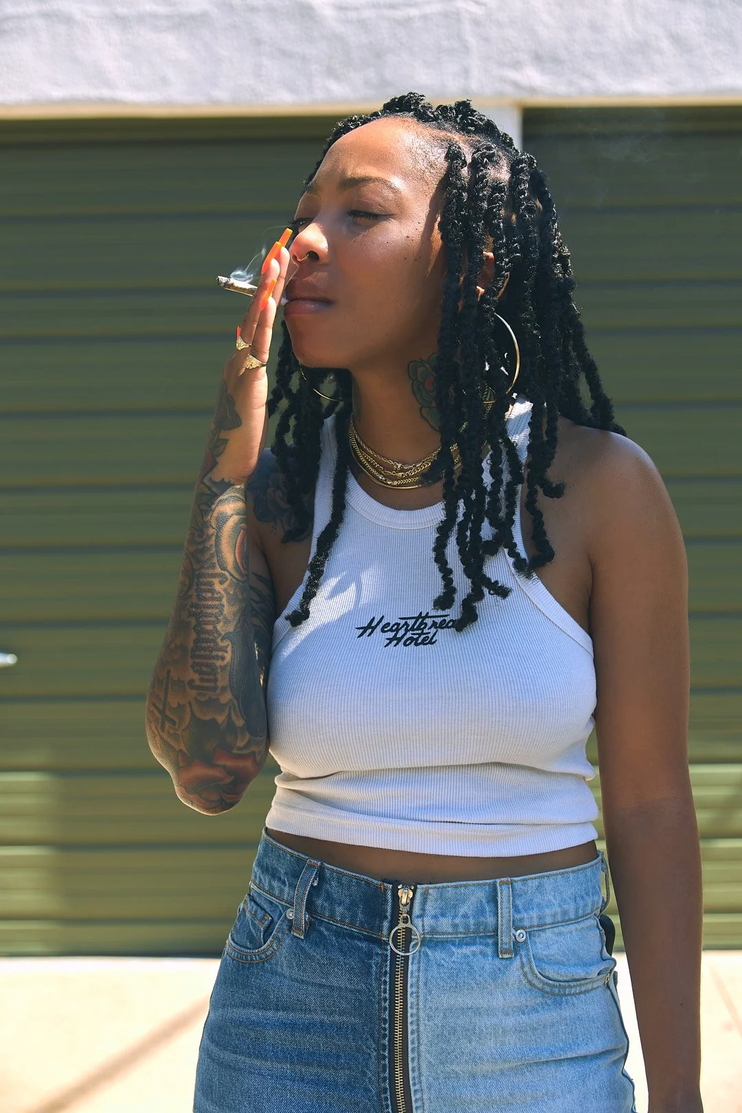 A woman with dreadlocks and tattoos is smoking a cigarette outdoors, wearing a white tank top and blue jeans.