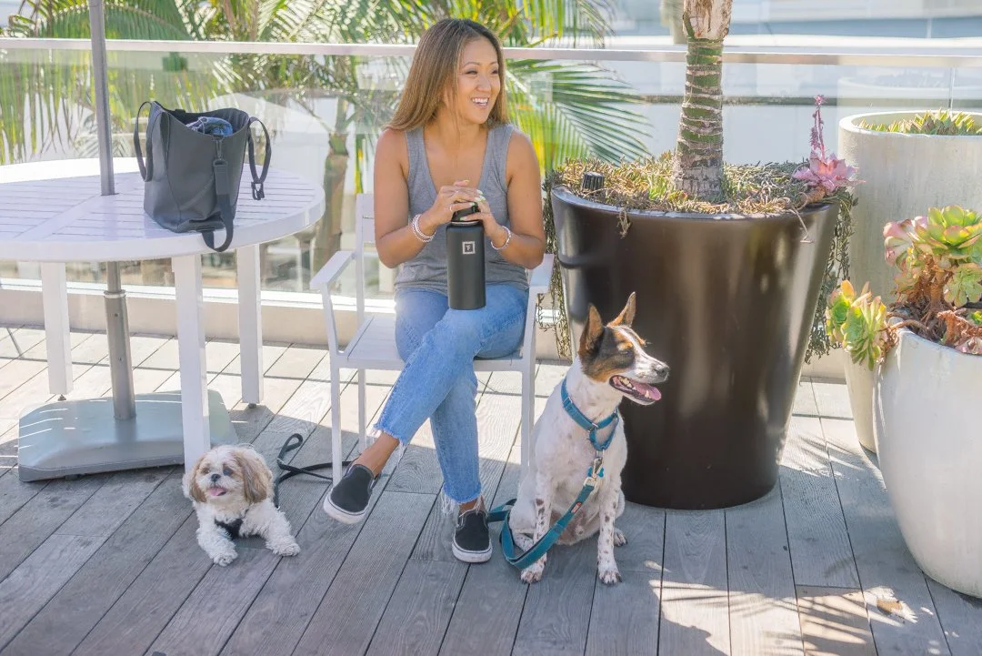 A woman sitting outside on a wooden deck with two dogs, a small white one and a larger black and white one, near potted plants and a glass railing.