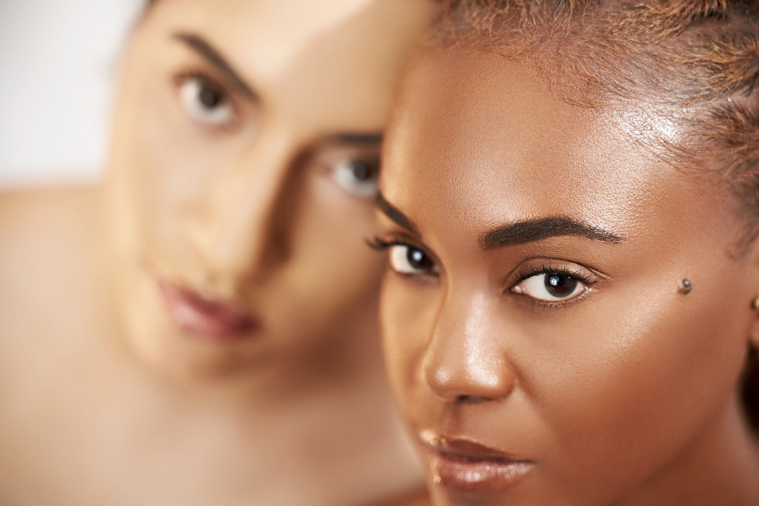 Close-up of two women with flawless skin, one with dark brown hair and the other with black hair, both with makeup and decorative piercings near their eyes, looking directly at the camera.