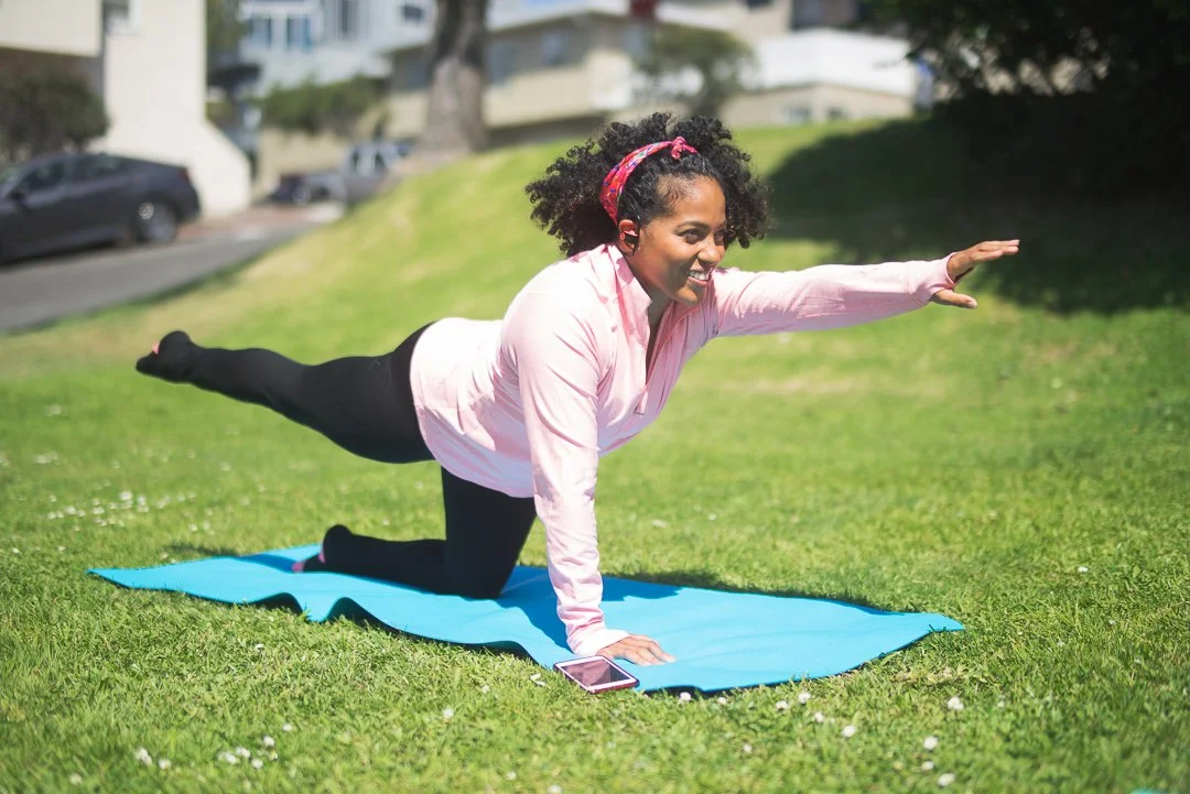 A woman practicing yoga outdoors on a grassy field, wearing a pink shirt, black leggings, and a pink headband, balancing on her hands with one arm extended forward, smiling, with a smartphone on her yoga mat.