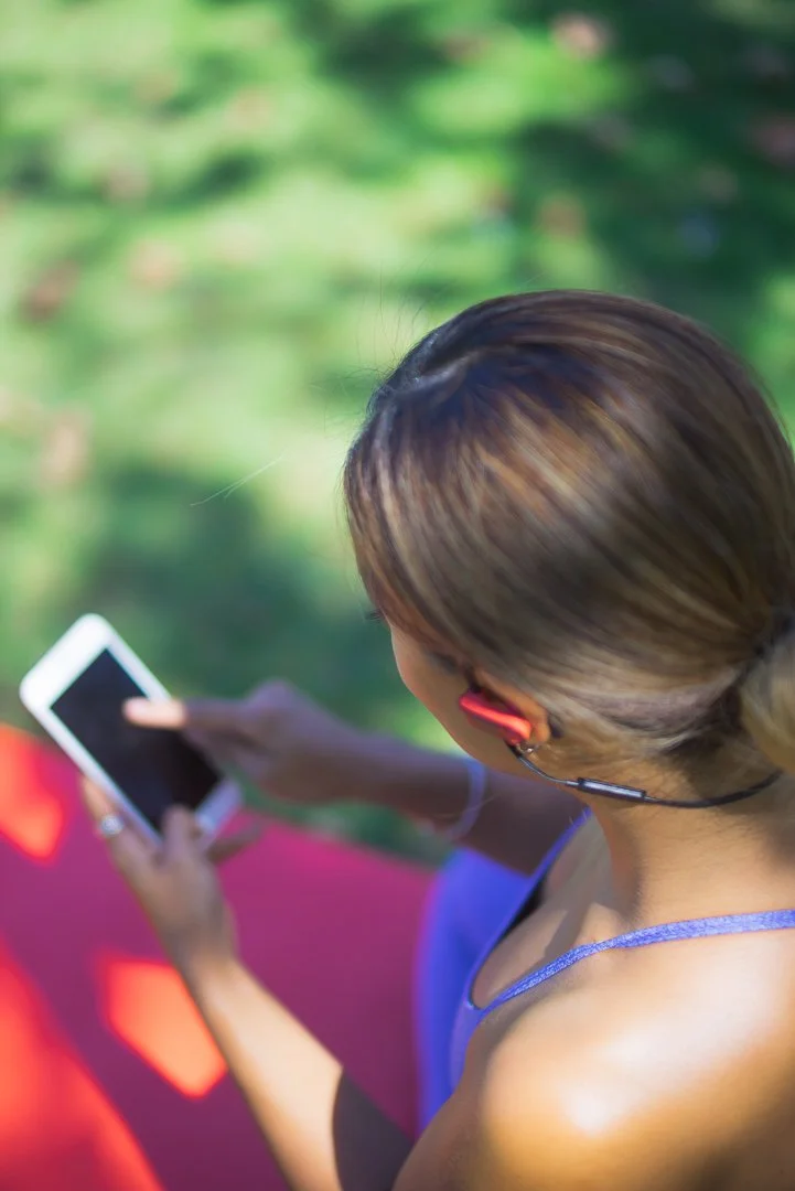 A woman exercising outdoors with a pink yoga mat, wearing headphones, holding a smartphone, with green foliage in the background.