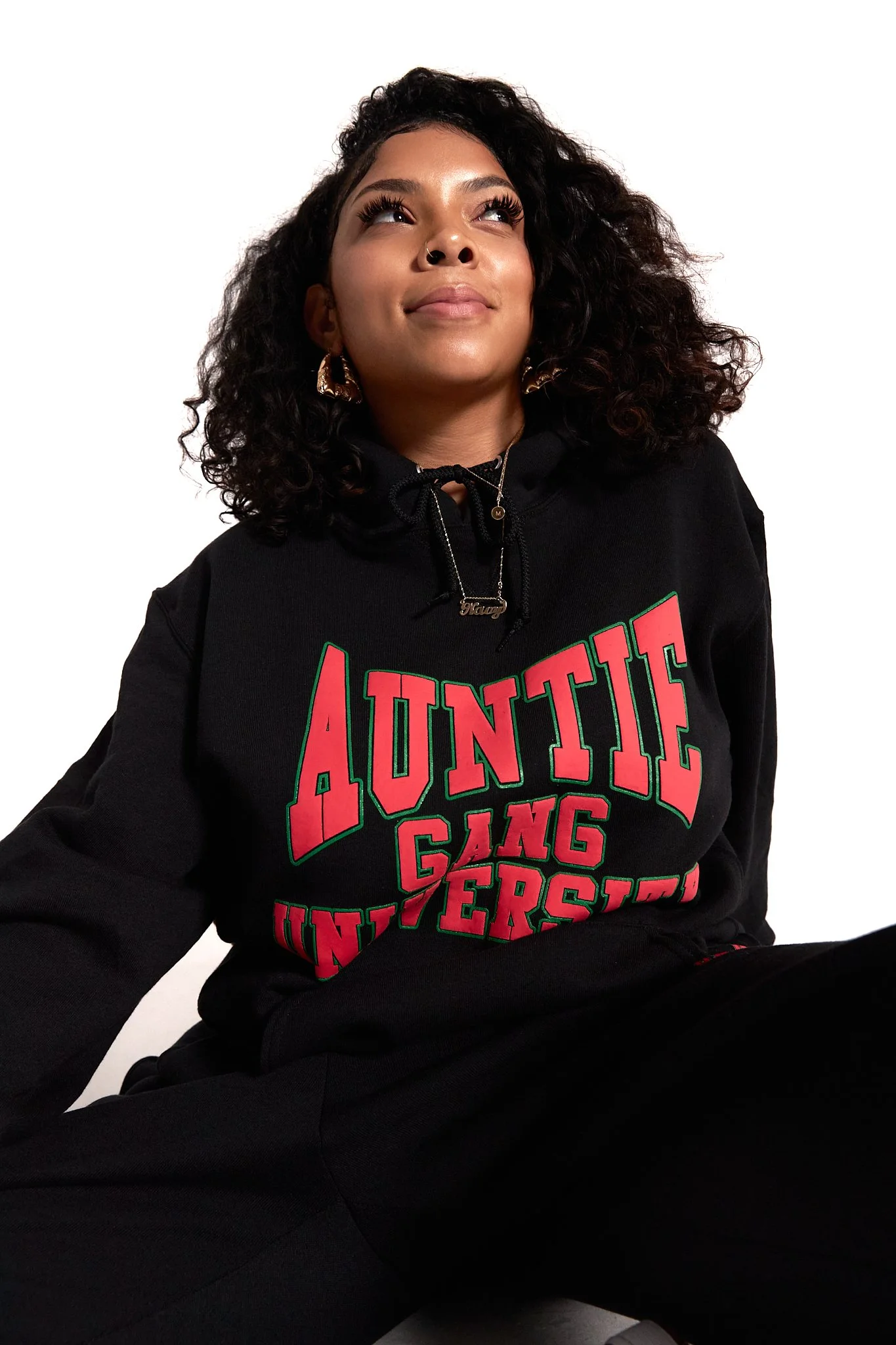A young woman with curly hair wearing a black hoodie with a bold pink and green graphic that reads 'Auntie Gang Universe' and black pants, sitting against a white background.