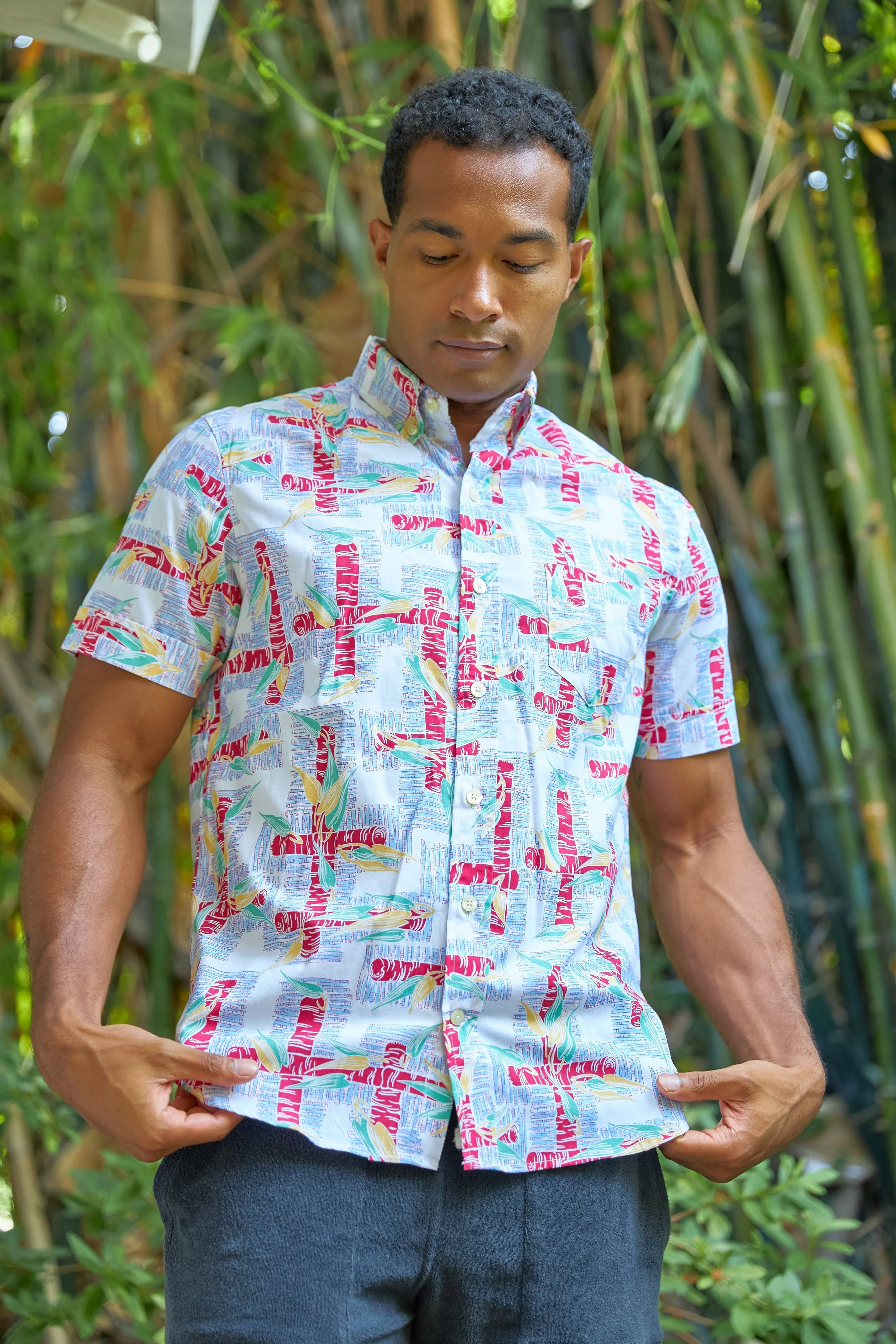 A man in a colorful, patterned short-sleeve shirt looking down, standing outdoors in a lush, green setting.